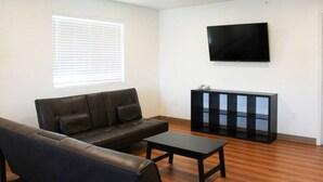 Ein Wohnzimmer mit einer Couch, einem Couchtisch, einem an der Wand befestigten Fernseher und einem Fenster mit Jalousien.