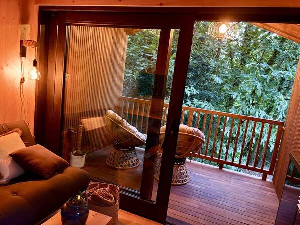 A wooden deck with a cushioned seating area, a glass door leading to a lush green outdoor space, and a small table with a vase.