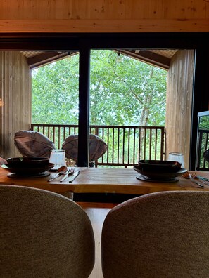 Een eetgedeelte met een tafel gedekt voor twee personen, uitzicht op bomen door grote ramen en een houten plafond.