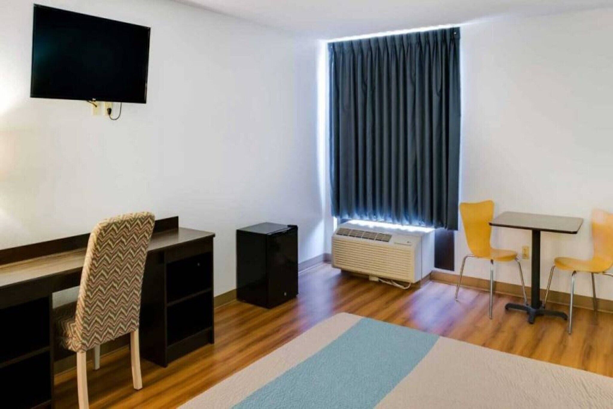 A room with a desk, chair, television, air conditioning unit, and a window with curtains.