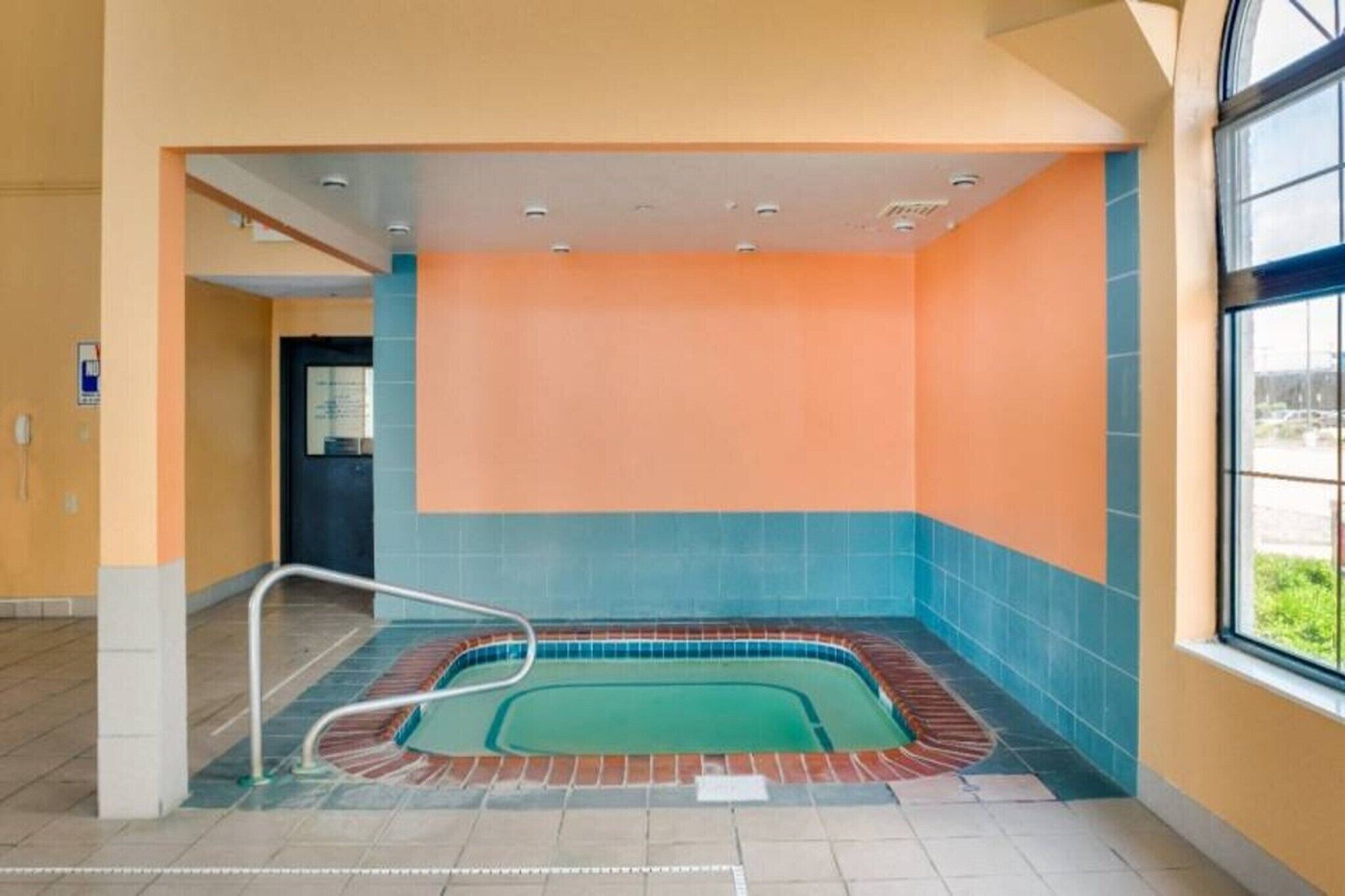 Una piscina con borde de azulejos, baranda metálica y vistas al exterior a través de una ventana.