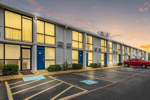 A row of motel rooms with individual doors, a parking lot, and a red vehicle parked in the lot.