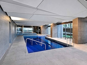 Una piscina interior moderna con paredes de vidrio transparente, un diseño de techo geométrico y suelo de baldosas.