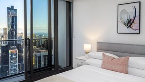 Et hotelværelse med udsigt over byen, en seng med hvide sengetæpper, en grå sengegavl og et natbord med en lampe.