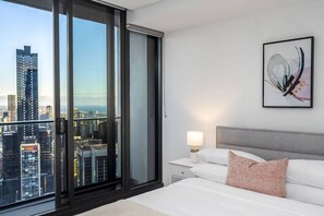 Ein Hotelzimmer mit Blick auf die Stadt, ein Bett mit weißer Bettwäsche, ein grauer Kopfteil und ein Nachttisch mit Lampe.