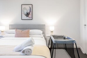 Une chambre d’hôtel équipée d’un lit, d’oreillers, d’une serviette roulée, d’un espace pour changer les bébés et d’une œuvre d’art abstraque encadrée accrochée au mur.