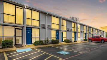 A row of motel rooms with individual doors, a parking lot, and a red vehicle parked in the lot.