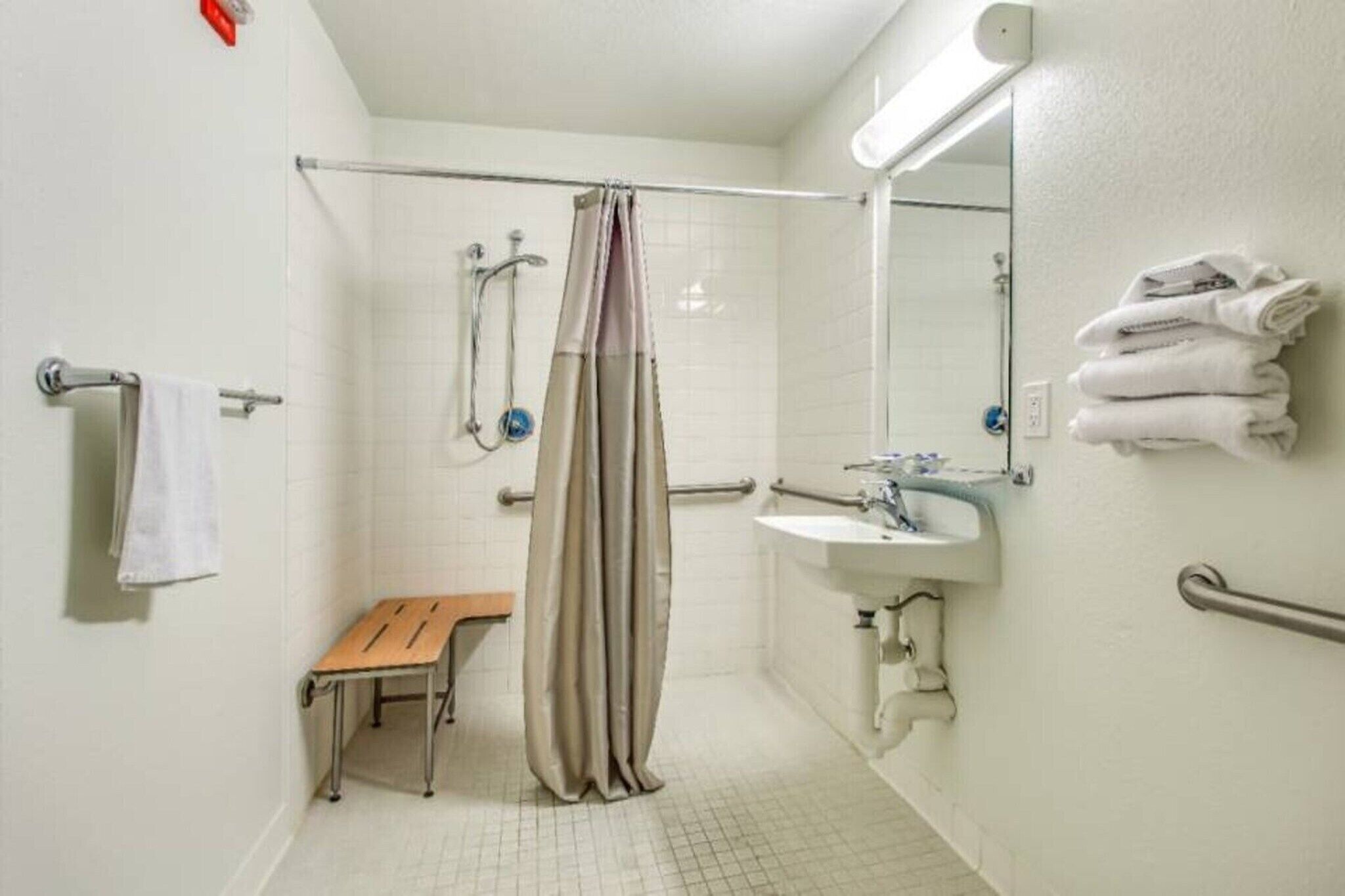 A bathroom with a shower, a sink, a mirror, a towel rack, and a bench.