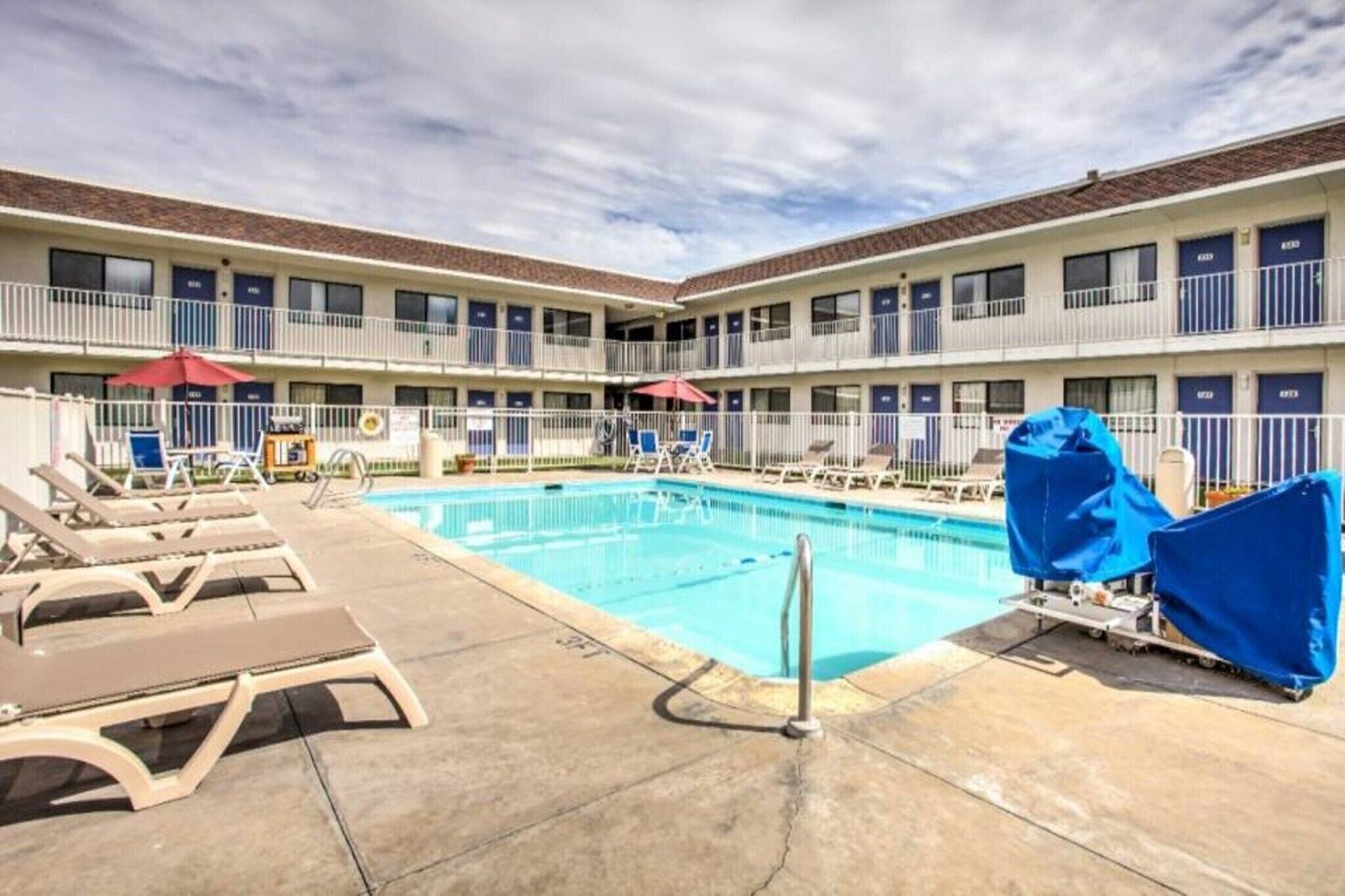 A hotel pool area with lounge chairs and umbrellas, surrounded by a multi-story building with balconies.