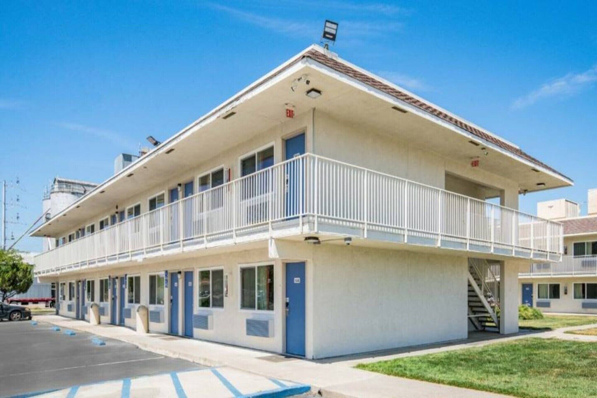 A two-story motel with a balcony on the upper floor, a parking lot, and a clear sky.