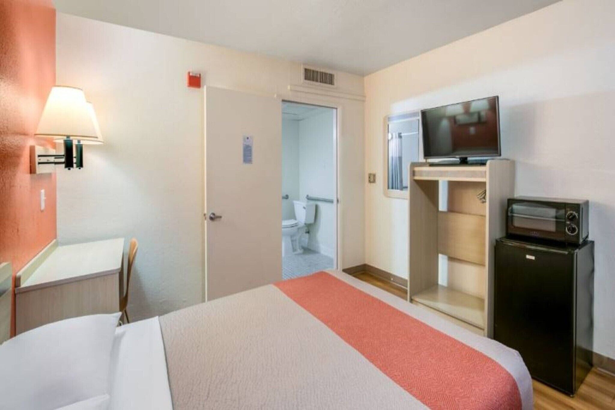 A hotel room with a bed, a television, a small refrigerator, and a bathroom visible through an open door.