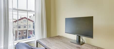 Una habitación con un escritorio de madera, una silla, un monitor de computadora y una ventana con vista a edificios.