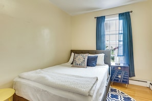 A bedroom with a bed, a nightstand, a window with curtains, and a patterned rug. - Walk to HBLR & Liberty State Park: Jersey City Apt (Jersey City)
