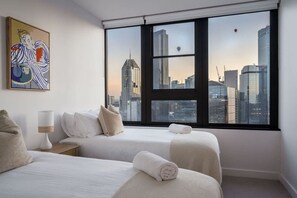 Ein Hotelzimmer mit zwei Betten, einem großen Fenster und Blick auf die Stadtlandschaft, einem Bild an der Wand und einer Lampe.