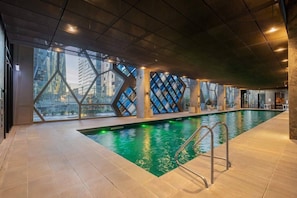An indoor swimming pool with a modern design, surrounded by large windows and geometric wall patterns. - Sky-high pool & sauna, near Marvel (Melbourne)