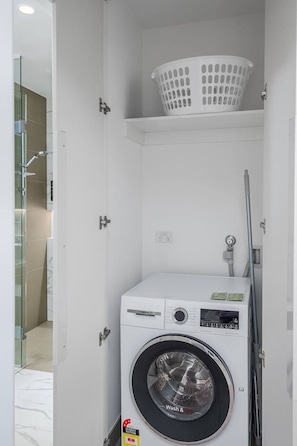 A compact laundry room with a front-loading washing machine, a white plastic basket on the shelf, and a glass shower enclosure. - Sky-high pool & sauna, near Marvel (Melbourne)