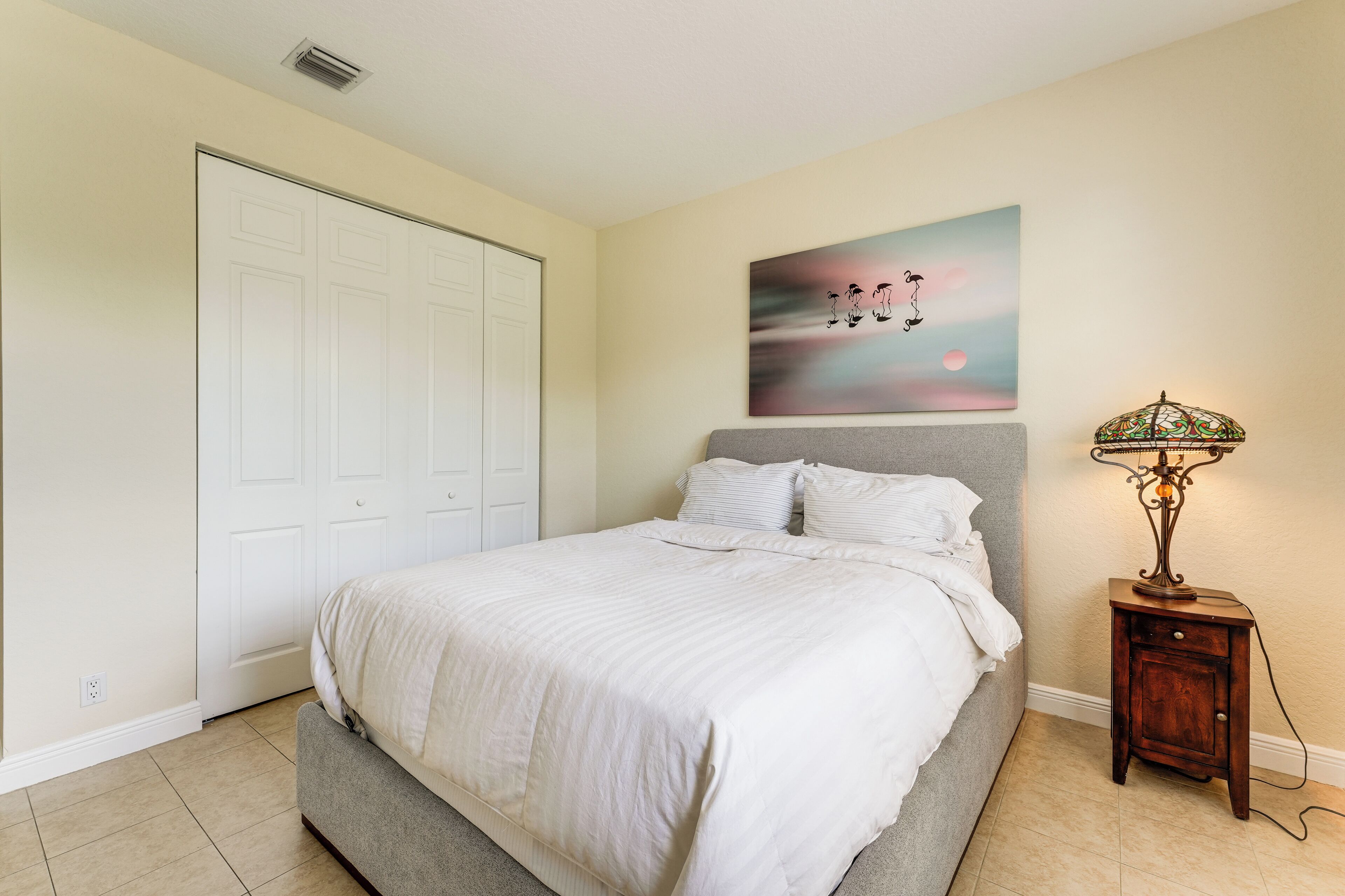 A bedroom with a bed, a nightstand, a lamp, a painting, and a closet.