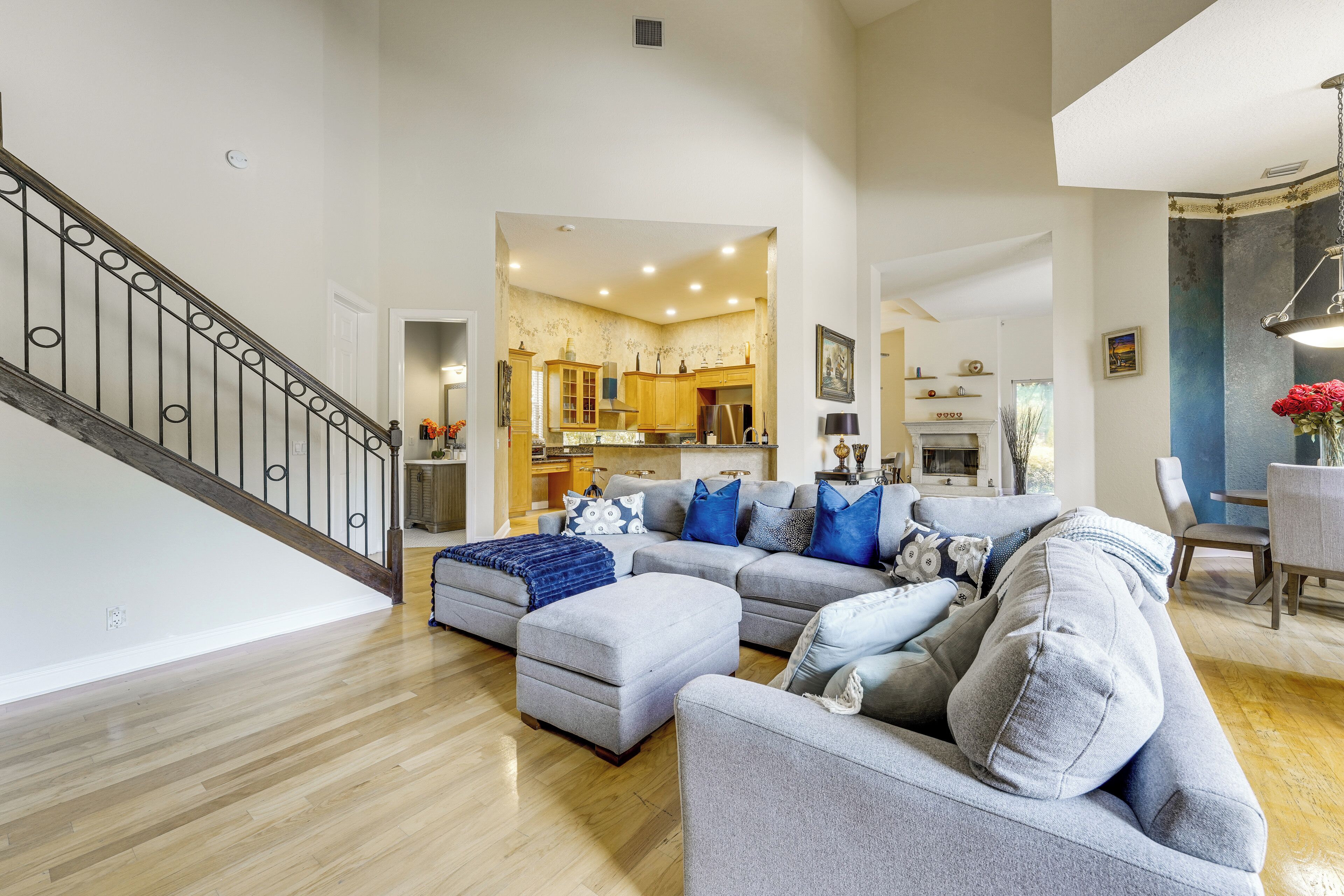 A spacious living room with a grey sectional sofa, a wooden staircase, and a kitchen in the background.