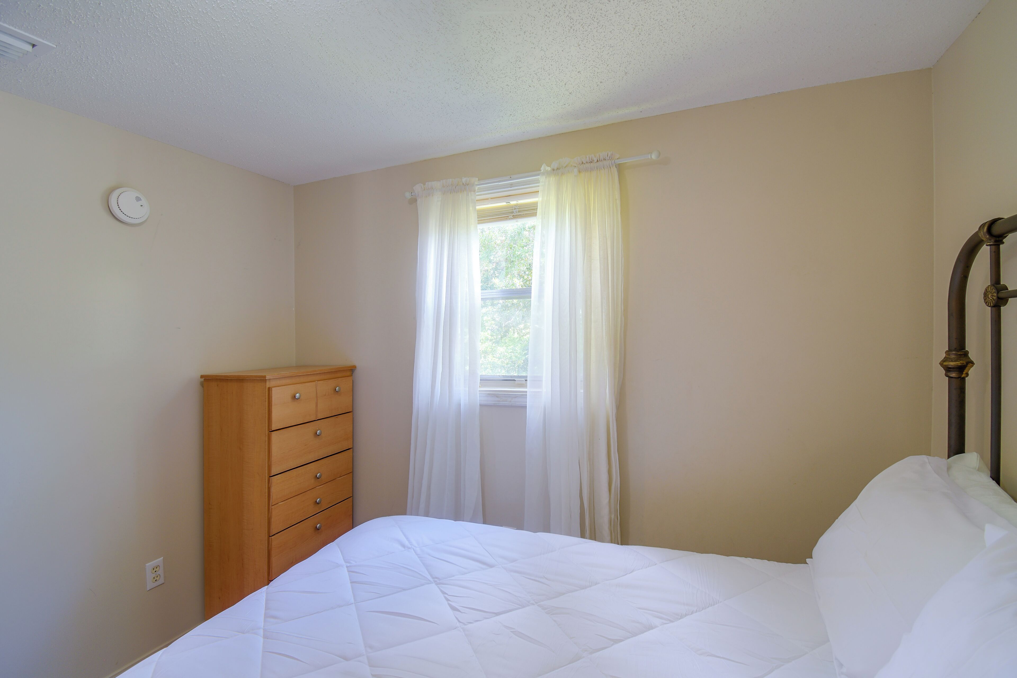 Un dormitorio con cama, cómoda de madera, ventana con cortinas y un interruptor de luz en la pared.