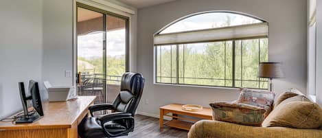 A modern office space with a desk, computer, chair, and a large window with a view of trees.