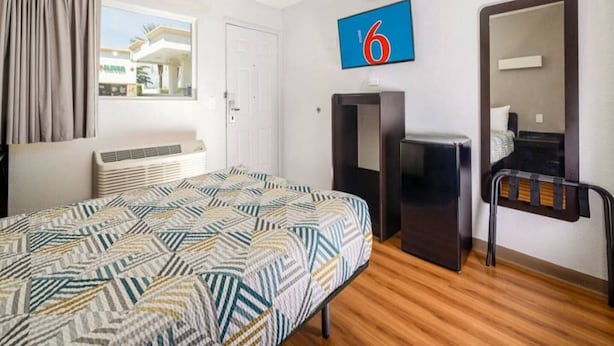 A hotel room with a bed, a desk, a chair, a mirror, and a window with curtains. - 1 QUEEN BED | NON-SMOKING, MINI FRIDGE (ONTARIO)