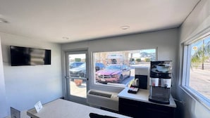 Una habitació amb escriptori, cafetera, televisor penjat a la paret i finestra amb vista a cotxes estacionats i un edifici a l'exterior.