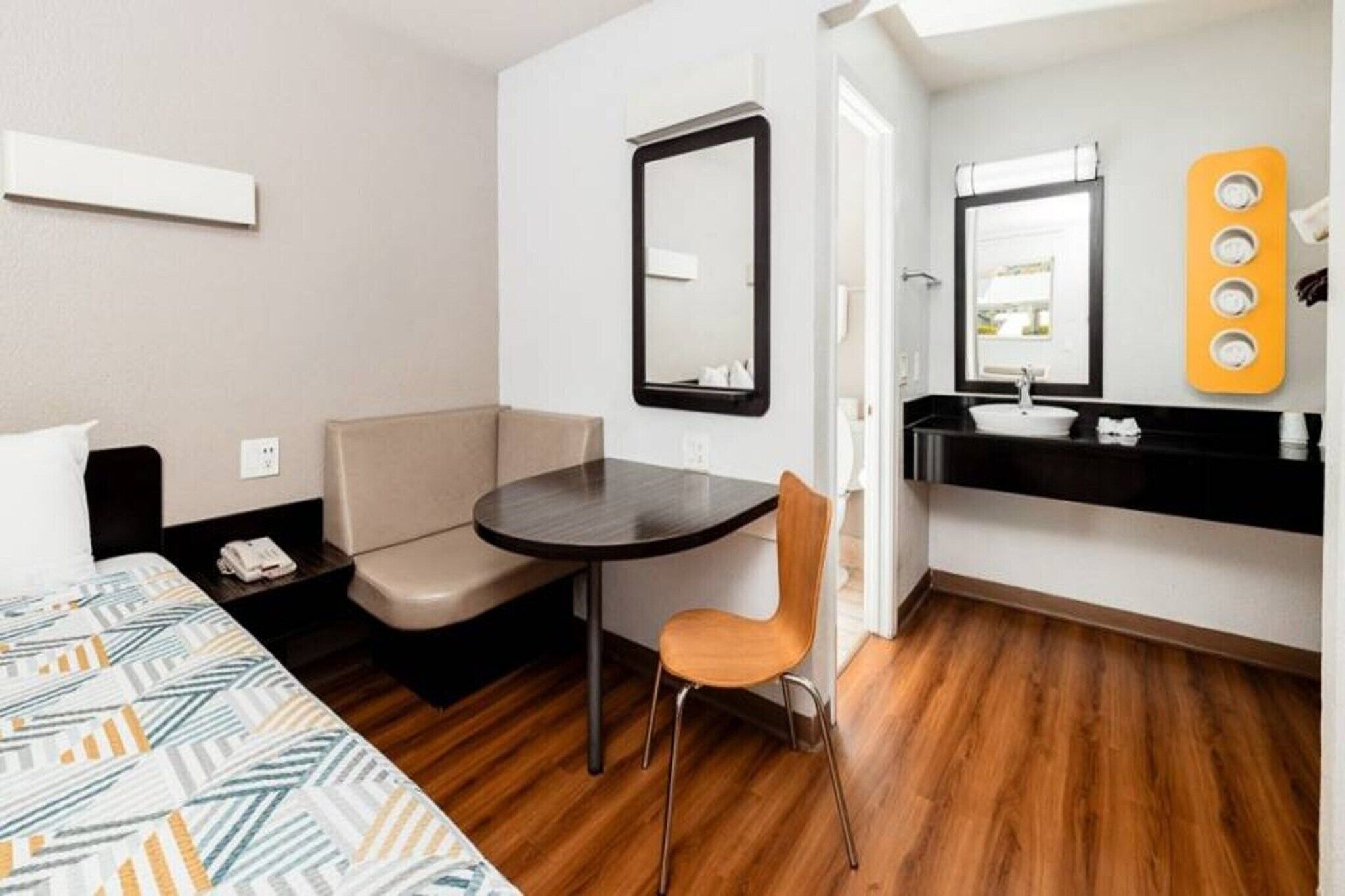 A compact hotel room with a bed, a small table, a chair, a sofa, a mirror, a sink, and a window.