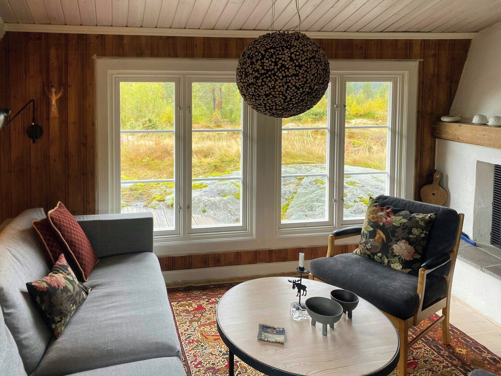 A cozy living room with a sofa, armchair, and round coffee table, large windows with a view of a natural landscape, and a hanging light fixture.