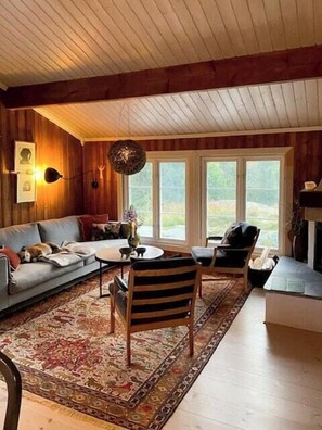 Ein gemütliches Wohnzimmer mit einer Couch, einem Couchtisch und einem Teppich. Große Fenster und ein Kamin sind vorhanden.