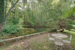 Ein friedlicher Außenbereich mit Metallstühlen und einem Tisch, umgeben von üppigem Grün und einer ruhigen Wasseroberfläche.
