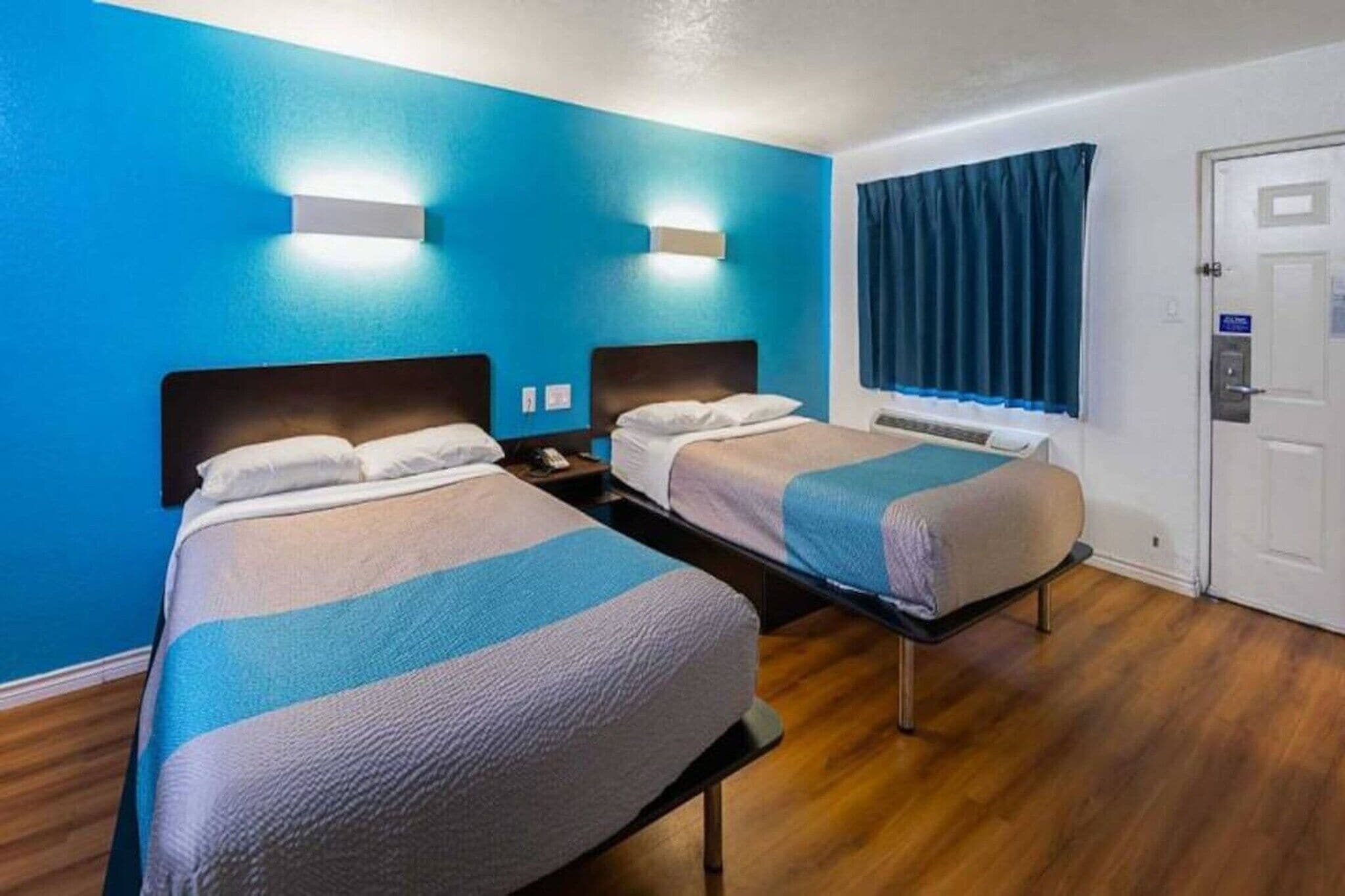 A hotel room with two beds, a blue wall, a window with curtains, and a door.