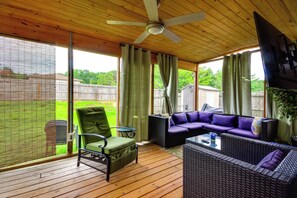Un espacio de estar techado al aire libre con ventilador de techo, una silla verde, un sofá con cojines morados y una mesa de centro de vidrio.