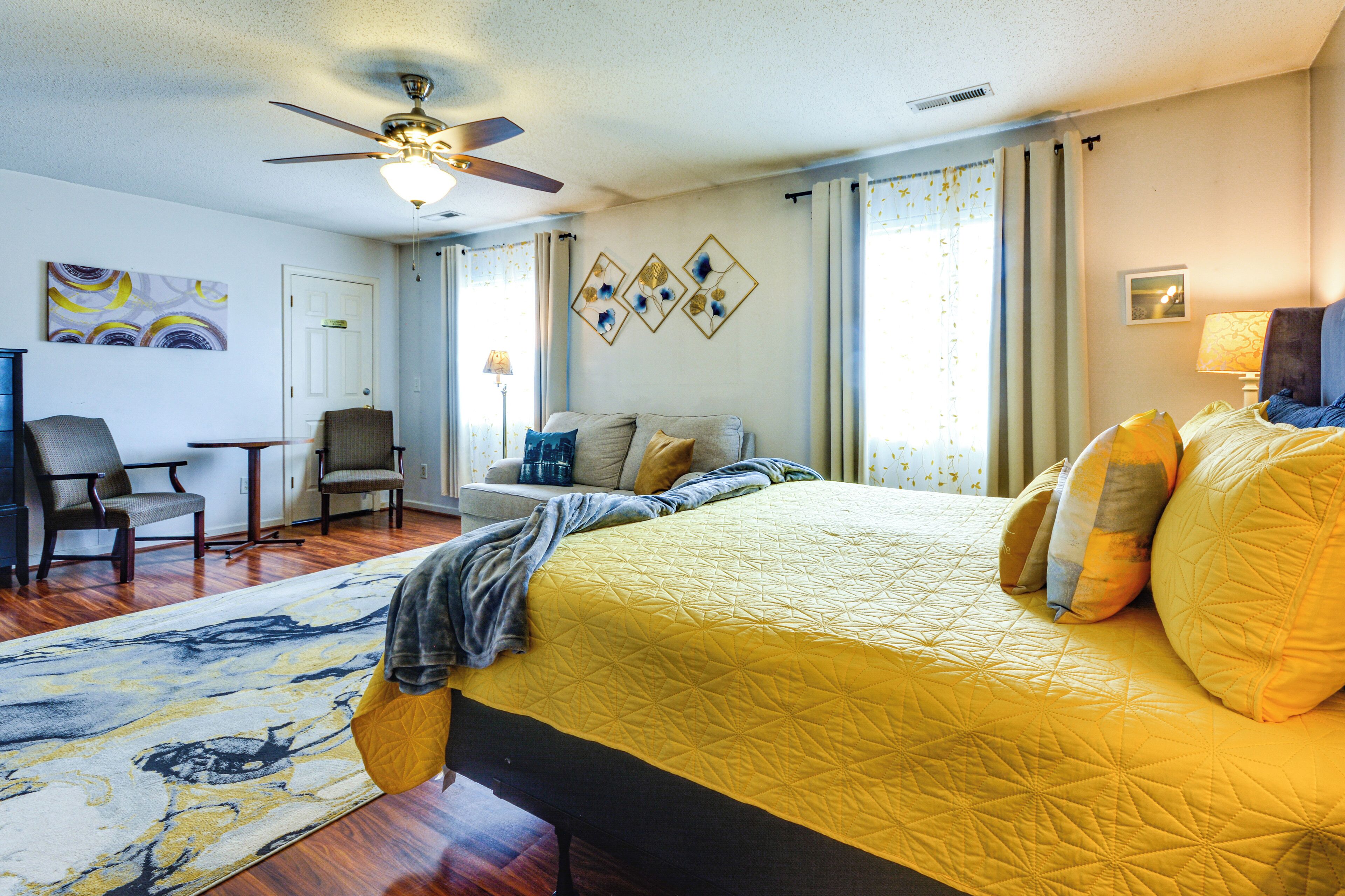 A hotel room with a bed, a desk, a chair, a sofa, a ceiling fan, and a window with curtains.