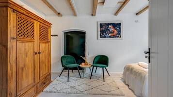 A room with a wooden wardrobe, two green chairs, a small table, a painting, and a bed.