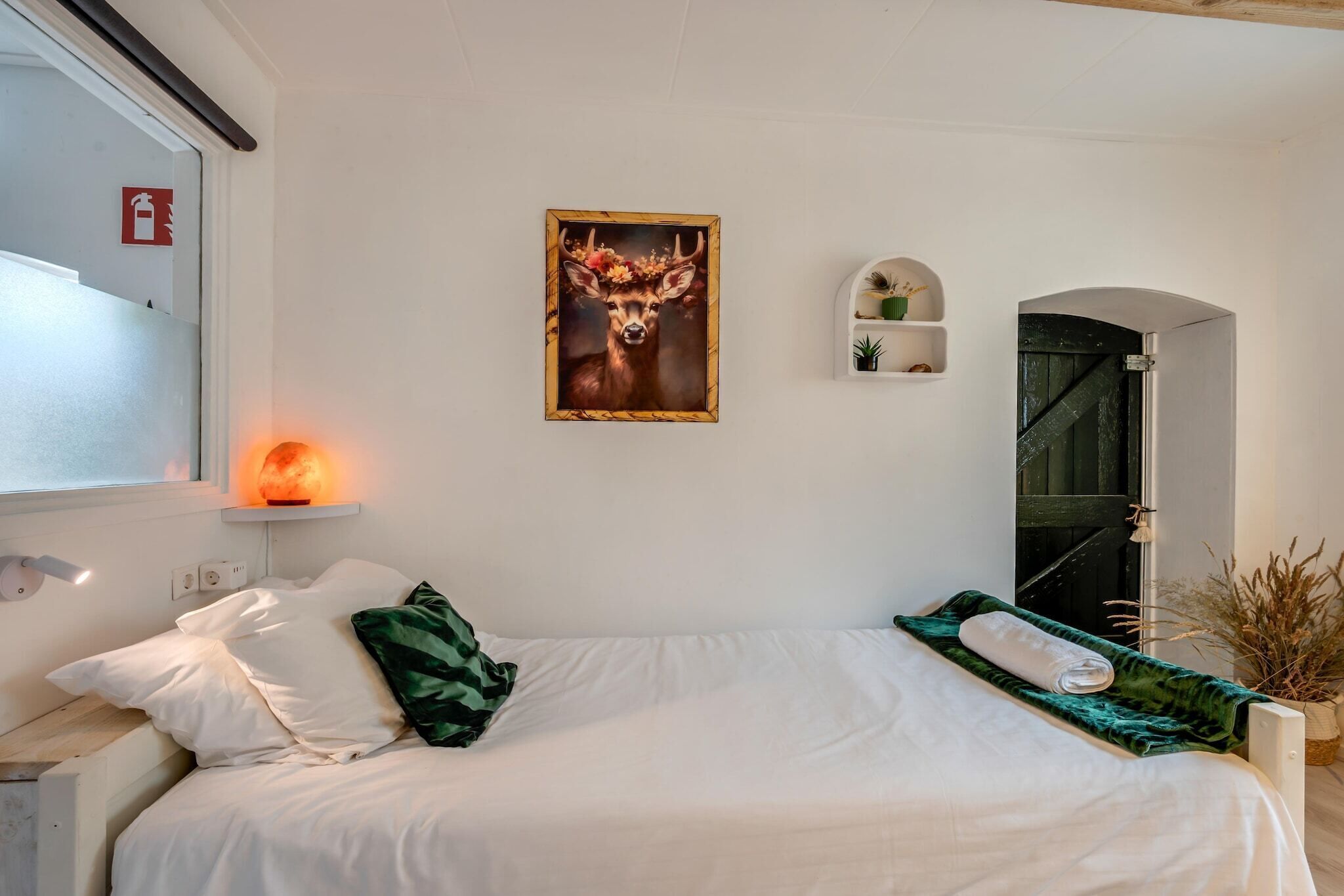 Une chambre à coucher avec un lit, des oreillers, un radiateur fixé au mur, un tableau encadré, une petite étagère avec des plantes et une porte donnant sur une autre pièce.