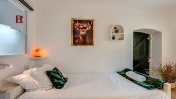 Une chambre à coucher avec un lit, des oreillers, un radiateur fixé au mur, un tableau encadré, une petite étagère avec des plantes et une porte donnant sur une autre pièce.
