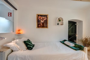 Une chambre à coucher avec un lit, des oreillers, un radiateur fixé au mur, un tableau encadré, une petite étagère avec des plantes et une porte donnant sur une autre pièce.