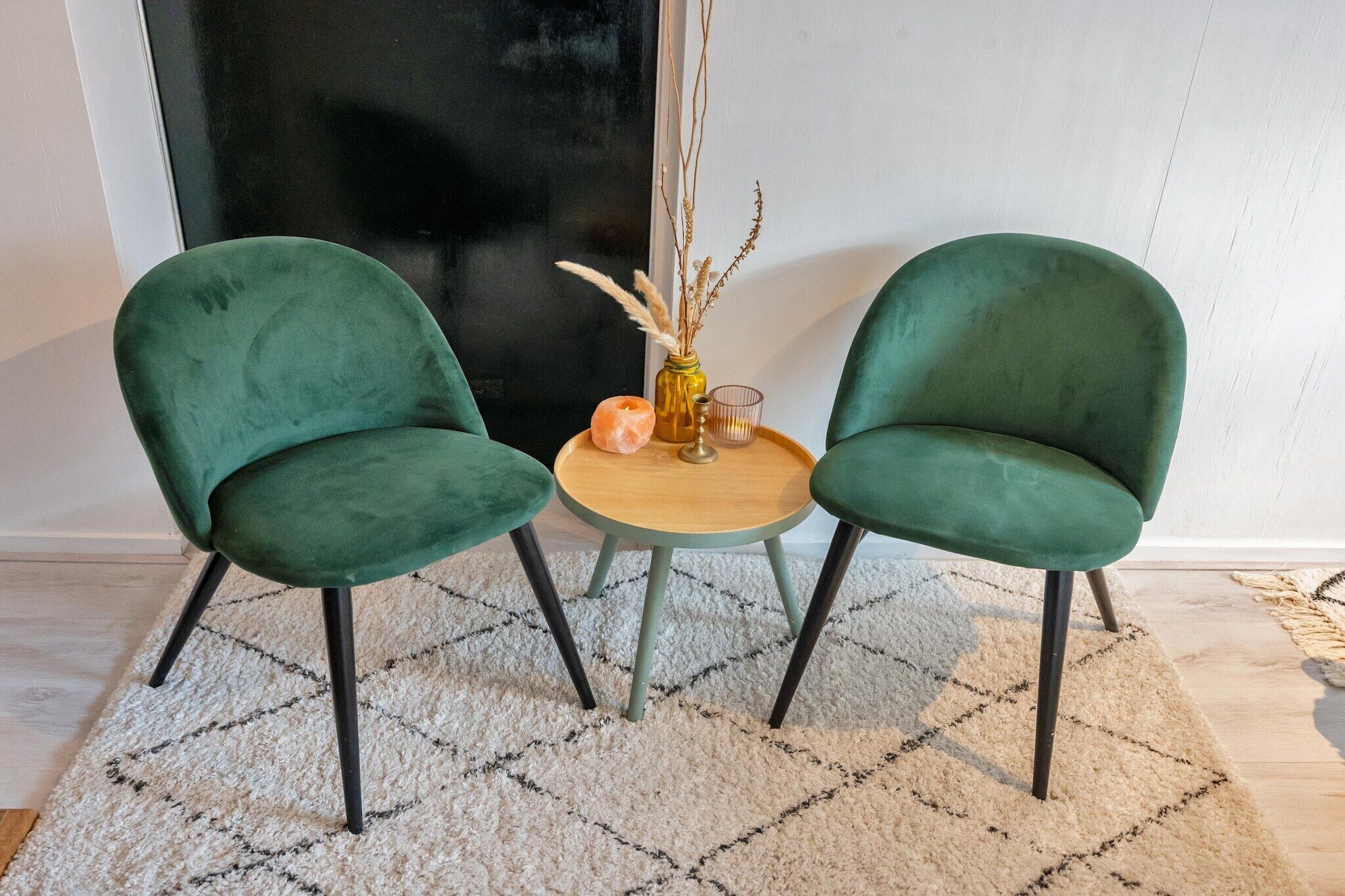 Deux fauteuils verts à revêtement en tissu, avec des pieds noirs, une petite table ronde en bois et un vase contenant des plantes séchées.