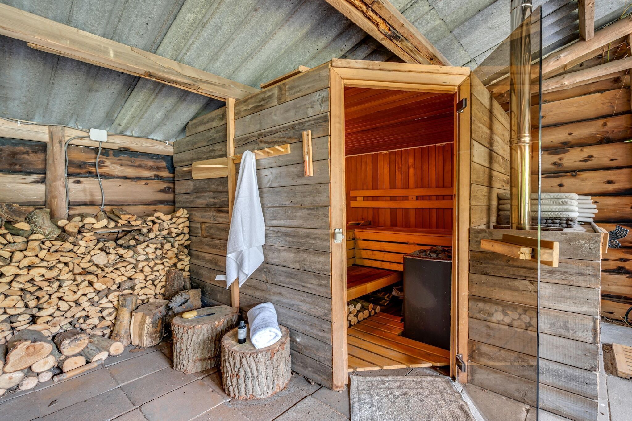 A sauna room with wooden walls and benches, a towel hanging on a rack, and a stack of firewood outside.