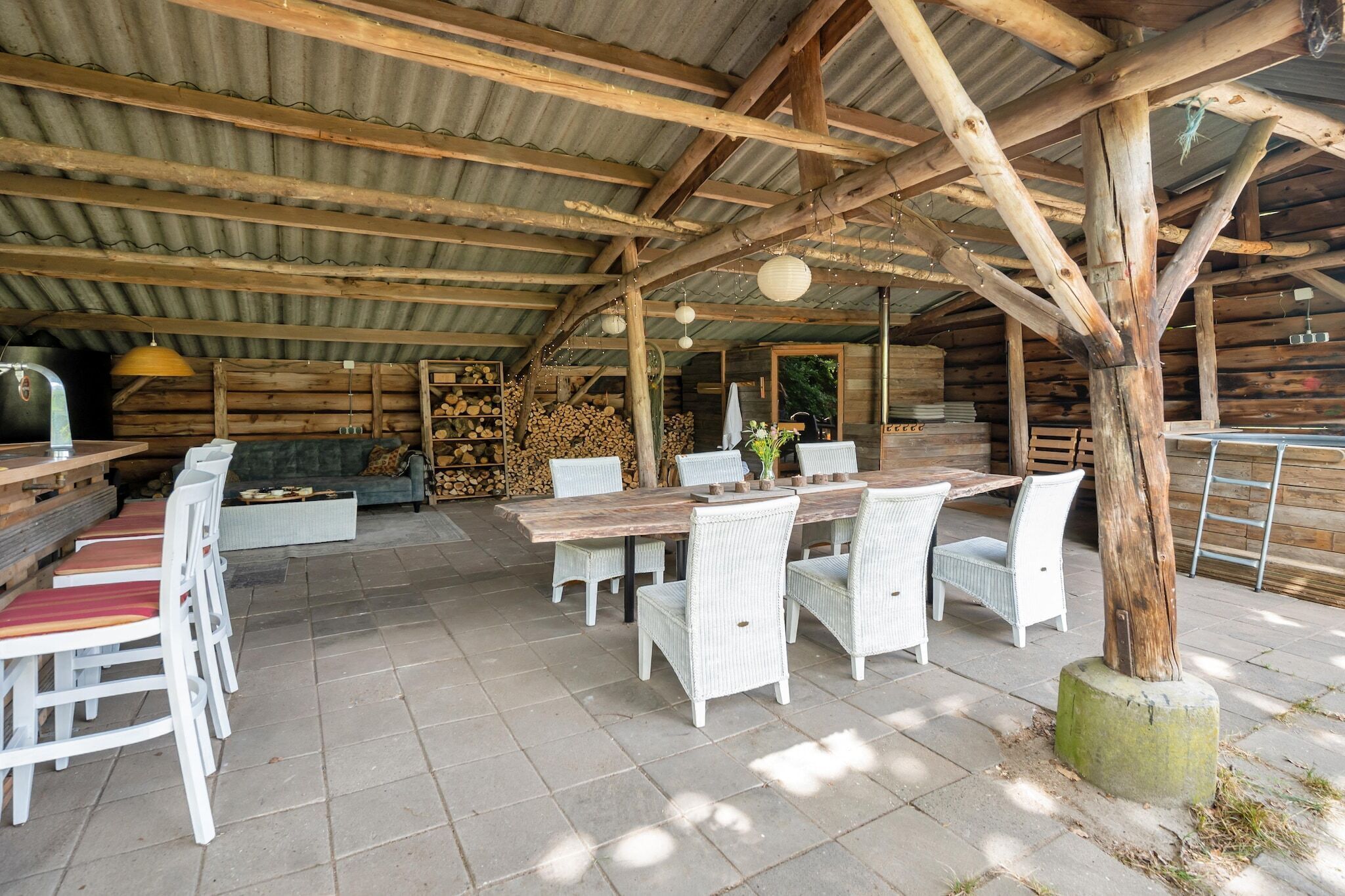 A rustic outdoor dining area with a wooden table and white wicker chairs, surrounded by a corrugated metal roof.