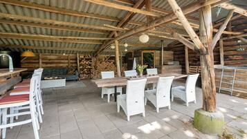 A rustic outdoor dining area with a wooden table and white wicker chairs, surrounded by a corrugated metal roof.