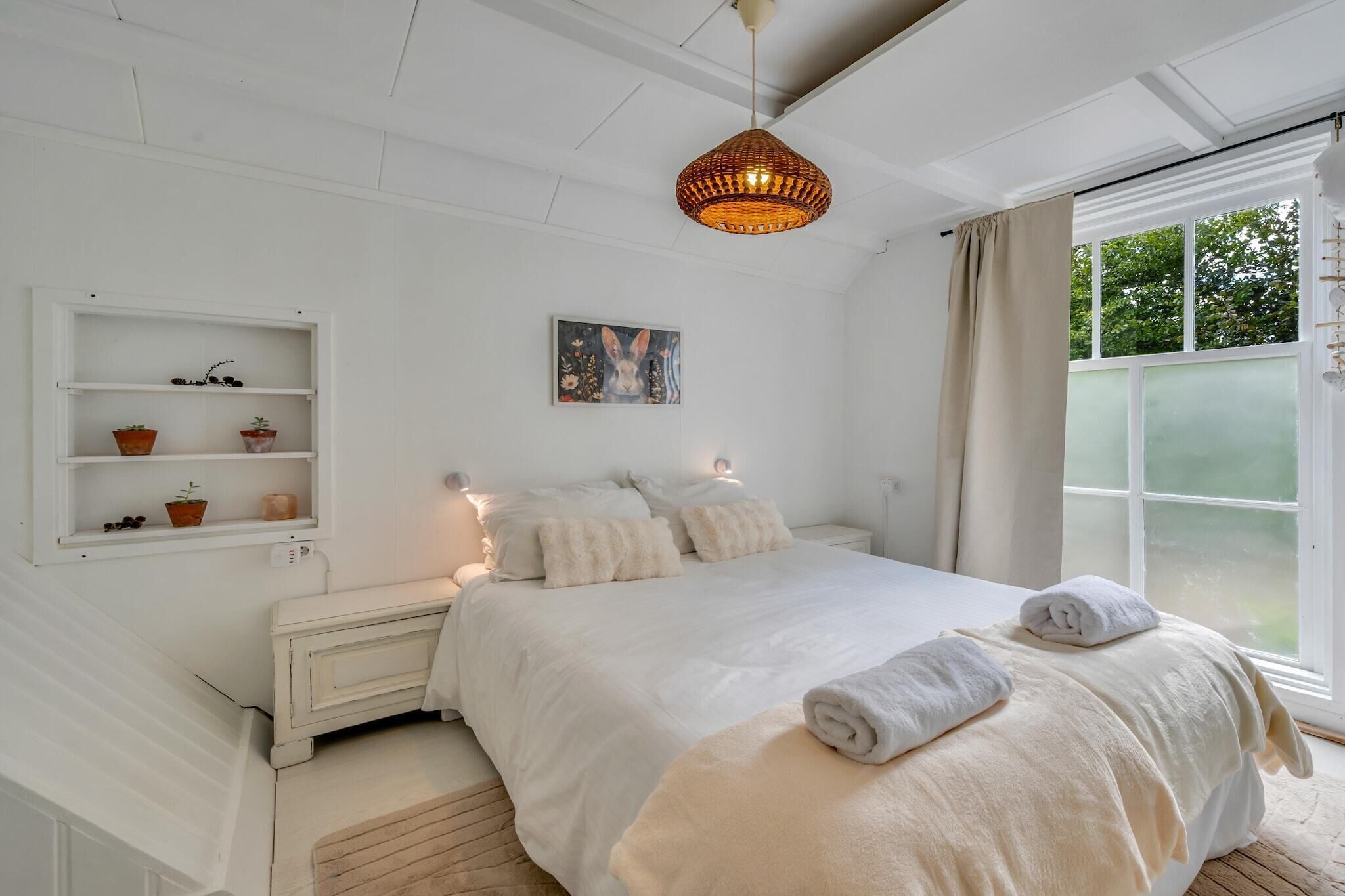 A bedroom with a bed, a bedside table, a shelf with plants, a painting, and a window with curtains.