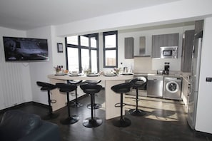A modern kitchen with a dining area, bar stools, a washing machine, and a TV mounted on the wall.
