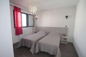 A bedroom with two beds, a red curtain, a wall-mounted light fixture, and a nightstand with a flower vase.