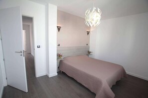 A bedroom with a bed, a door, a nightstand, a wall-mounted light fixture, and a small table with a vase of flowers. - Villa in Anglet between ocean, forests (Anglet)