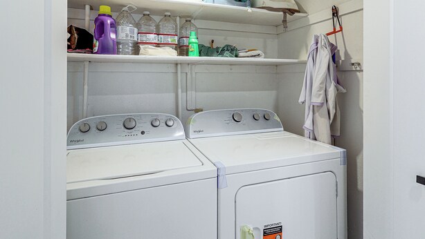 A laundry room with two Whirlpool washers, shelves above containing cleaning supplies, and a hanging rack with towels. - Colorado River Family Escape in Bullhead City (Bullhead City)