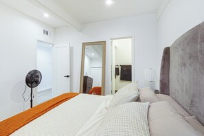Ein Schlafzimmer mit einem Bett, einem Nachttisch mit Lampe, einem Spiegel und einem Standventilator.