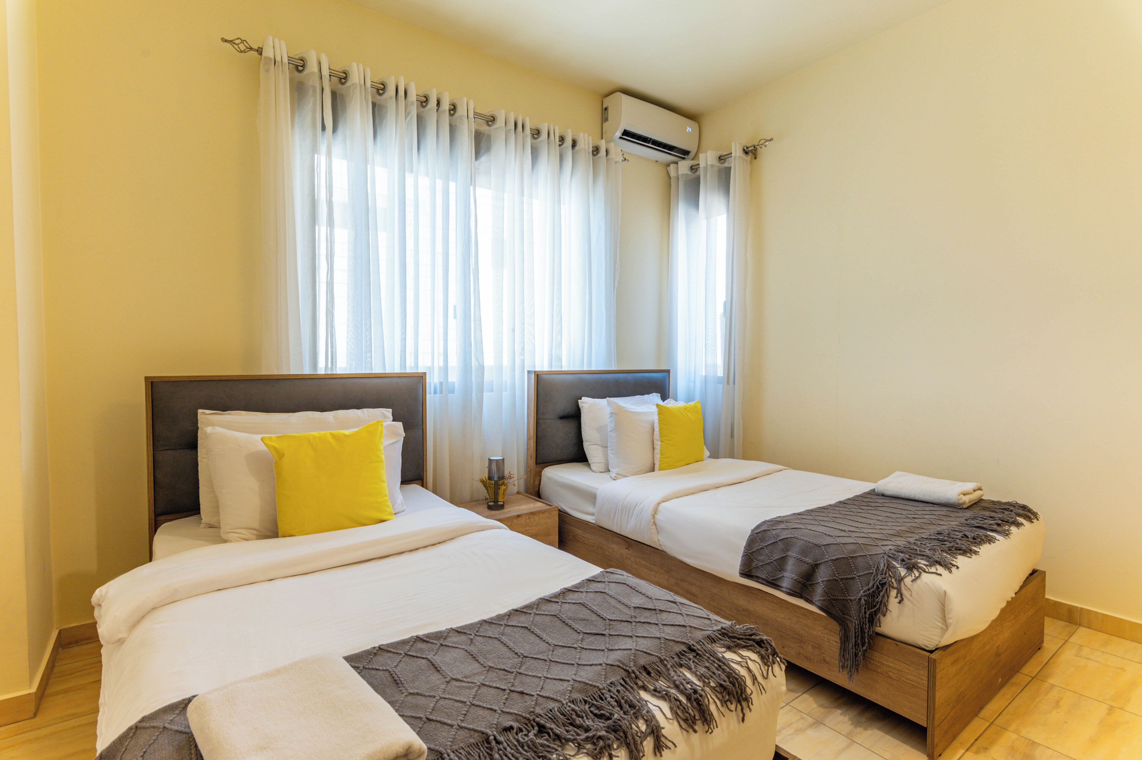 A hotel room with two beds, a window with sheer curtains, an air conditioning unit, and a small bedside table with a lamp.
