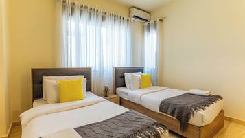 A hotel room with two beds, a window with sheer curtains, an air conditioning unit, and a small bedside table with a lamp.
