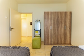 Quarto de hotel com duas camas, um guarda-roupa de madeira, um banquinho verde e um espelho.